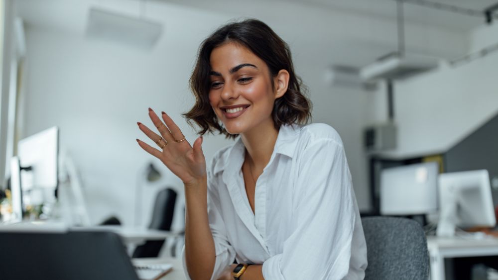 Mujer con camisa blanca en una oficina saludando a su portátil