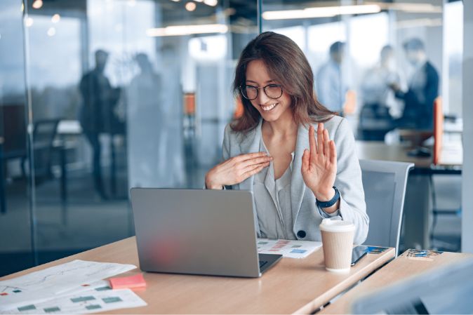 Bir ofiste dizüstü bilgisayarının önünde oturan kadın, çevrimiçi toplantıda bir şey söylemek istediğine dair elini kaldırıyor
